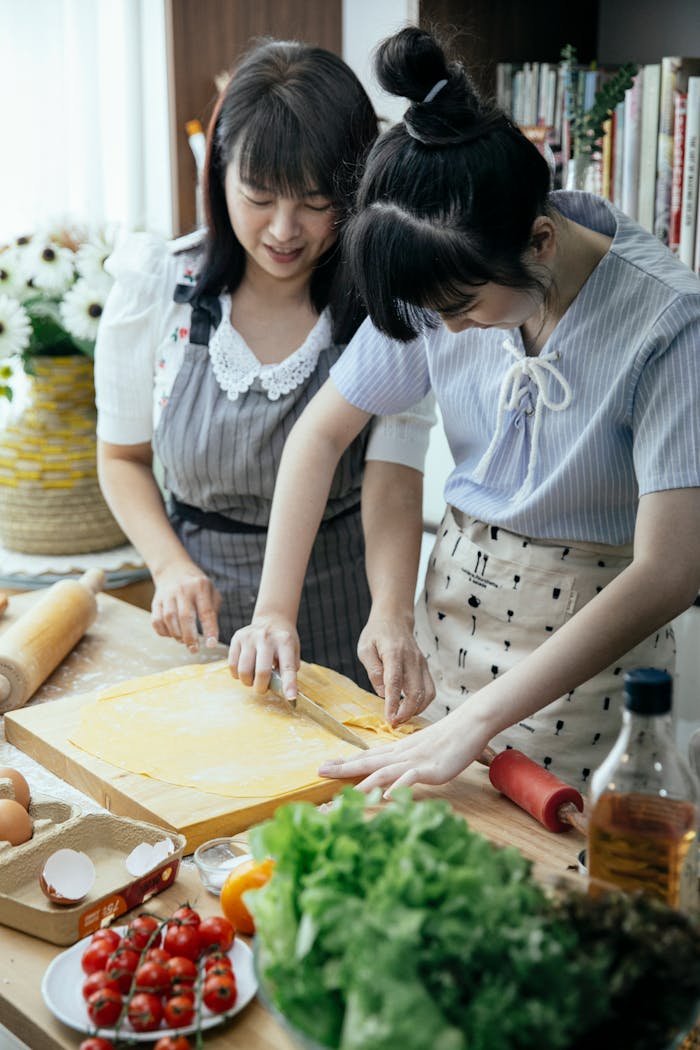 High angle of Asian women in aprons cutting dough for pasta on table with vegetables eggs and cooking ingredients