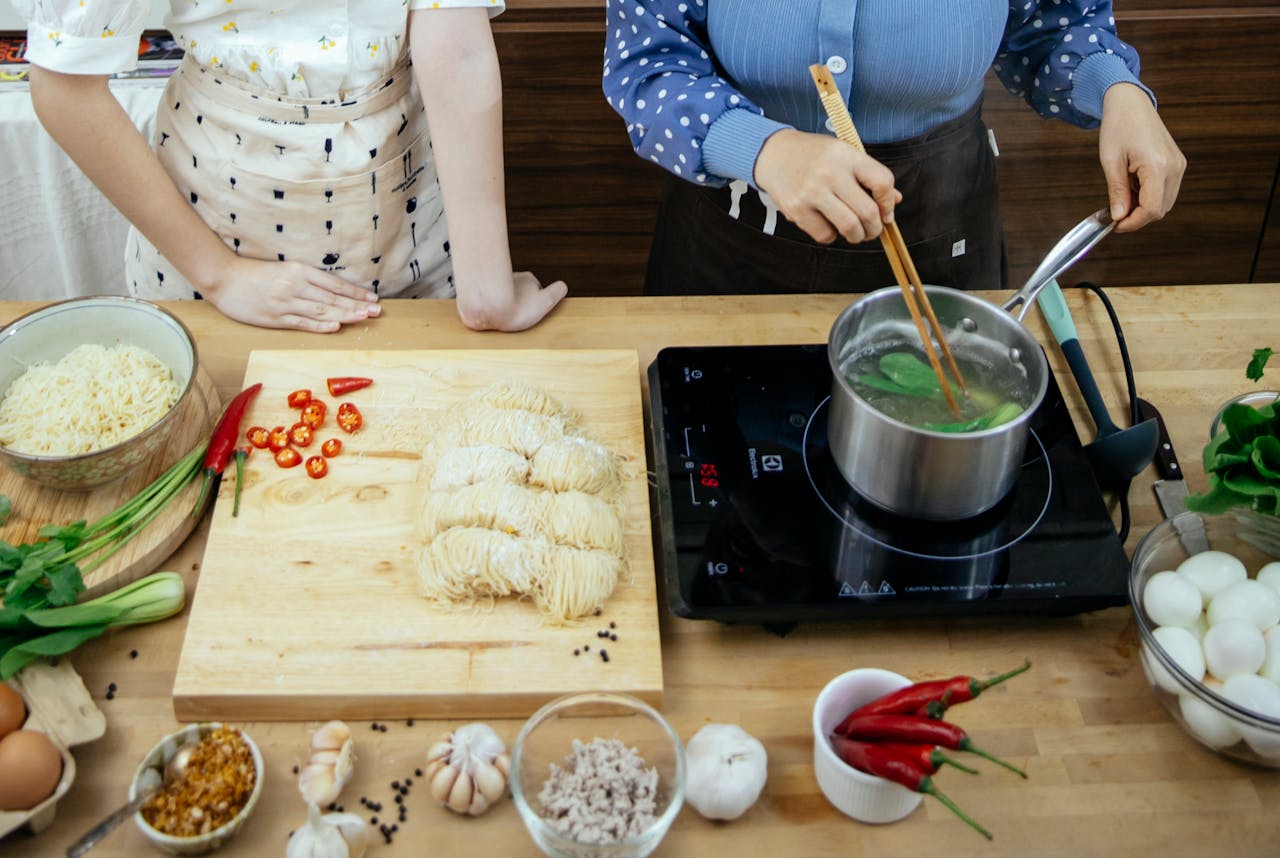 High angle of unrecognizable women boiling greens in saucepan making noodles with spices