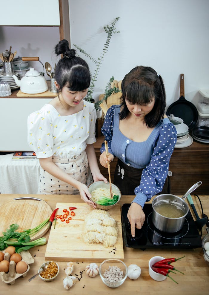 Two Asian women joyfully preparing a homemade noodle dish in a modern kitchen setting.